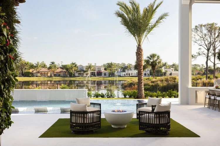 Luxury outdoor patio featuring modern lounge seating, fire pit, and pool with a scenic waterfront and palm tree backdrop
